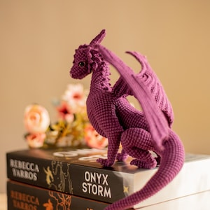 May include: A handcrafted, purple crochet dragon figurine with detailed wings, tail, and scales. The dragon is perched atop a stack of books with titles like "Onyx Storm" and "Fourth Wing". The background features soft-focus flowers.