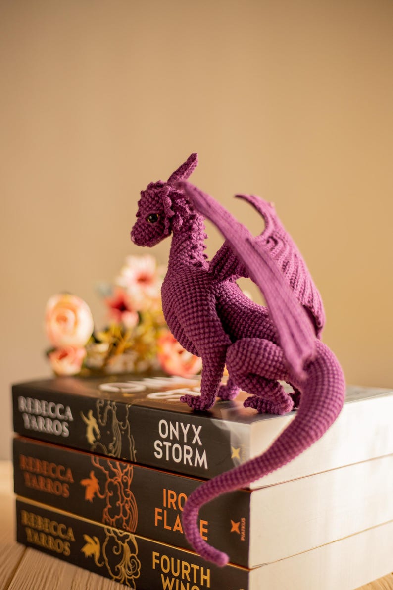 May include: A handcrafted, purple crochet dragon figurine with detailed wings, tail, and scales. The dragon is perched atop a stack of books with titles like "Onyx Storm" and "Fourth Wing". The background features soft-focus flowers.