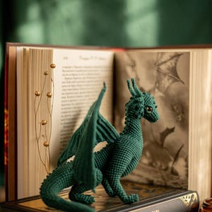 May include: A dark green crochet dragon figurine with detailed wings and a textured body. The dragon is posed on a decorative book with gold accents, in front of an open book with illustrations.