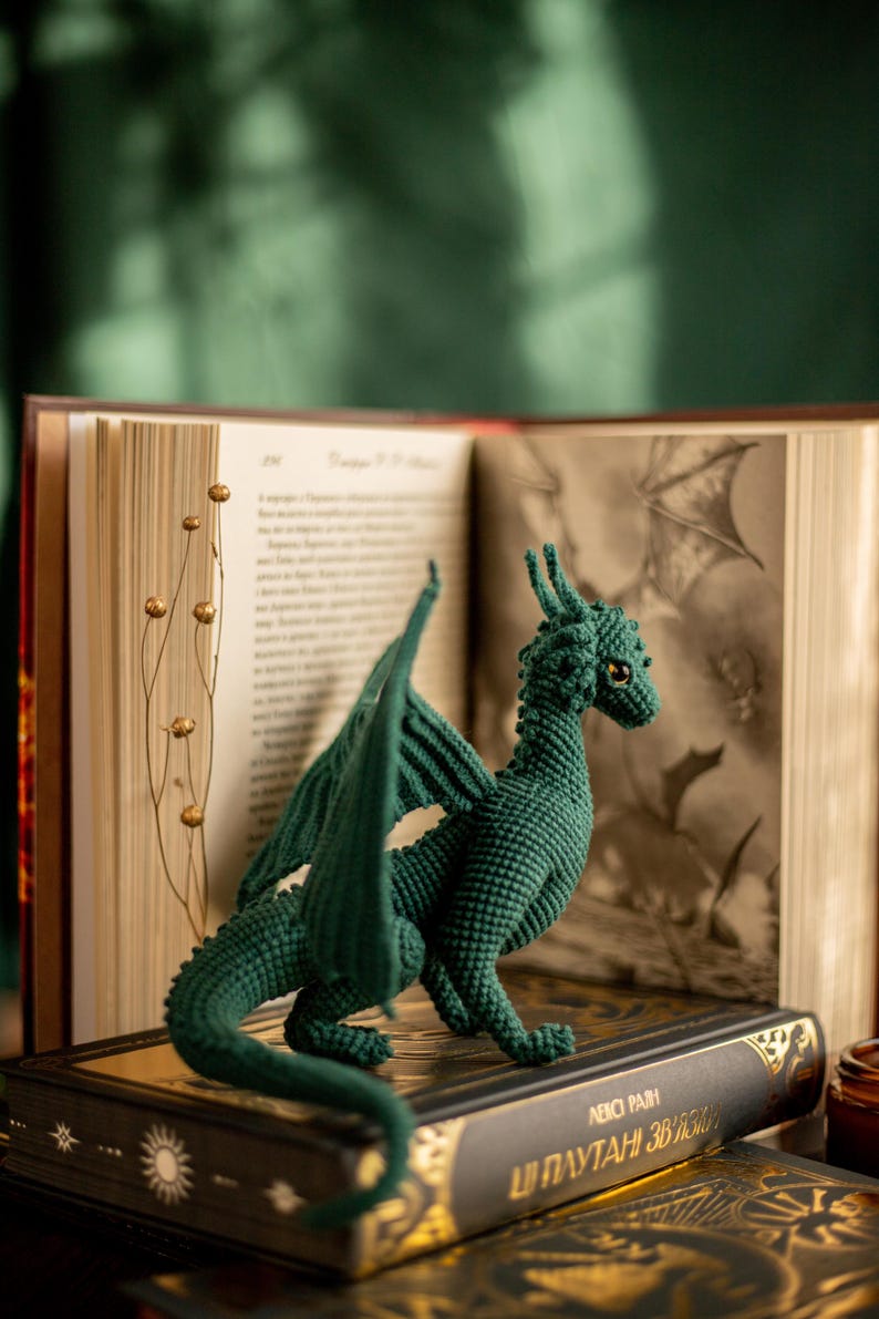 May include: A dark green crochet dragon figurine with detailed wings and a textured body. The dragon is posed on a decorative book with gold accents, in front of an open book with illustrations.