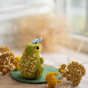 Puede incluir: Una rana verde hecha a mano con ganchillo, con detalles florales bordados y una peque&ntilde;a mariposa azul en la cabeza. La rana est&aacute; sentada sobre una estera verde, rodeada de flores amarillas secas. Un objeto decorativo encantador y caprichoso.