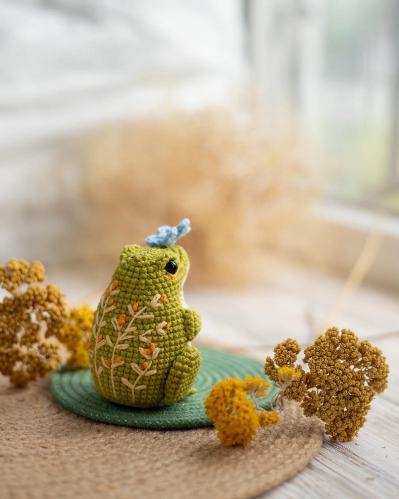 Puede incluir: Una rana verde hecha a mano con ganchillo, con detalles florales bordados y una peque&ntilde;a mariposa azul en la cabeza. La rana est&aacute; sentada sobre una estera verde, rodeada de flores amarillas secas. Un objeto decorativo encantador y caprichoso.