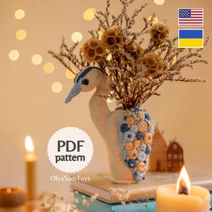 Op de afbeelding: Een handgemaakte gehaakte vogelvaas in crème, blauw en oranje. De vaas is gevuld met gedroogde bloemen en staat op boeken. De tekst "PDF pattern" is zichtbaar. Een uniek decoratie-item.