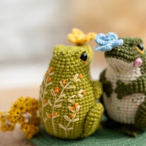 Puede incluir: Dos figuras de rana de ganchillo hechas a mano. Una rana es verde liso con detalles florales bordados, la otra es verde y blanca. Ambas ranas tienen peque&ntilde;os acentos florales y ojos negros. Est&aacute;n sobre una alfombra verde.