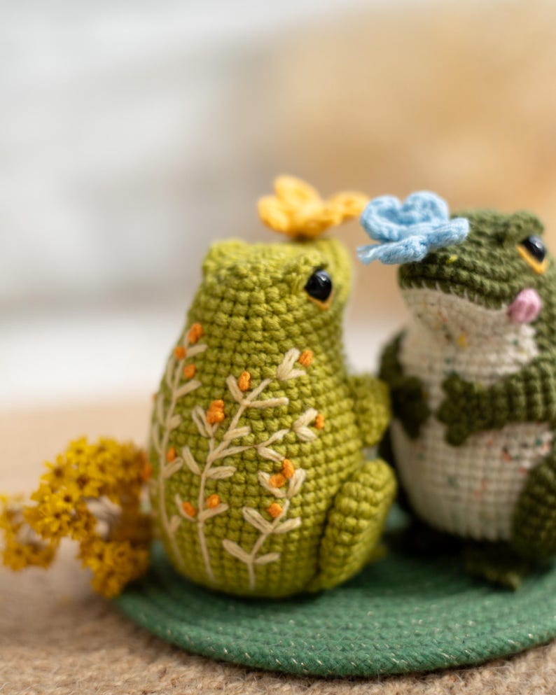 Puede incluir: Dos figuras de rana de ganchillo hechas a mano. Una rana es verde liso con detalles florales bordados, la otra es verde y blanca. Ambas ranas tienen peque&ntilde;os acentos florales y ojos negros. Est&aacute;n sobre una alfombra verde.