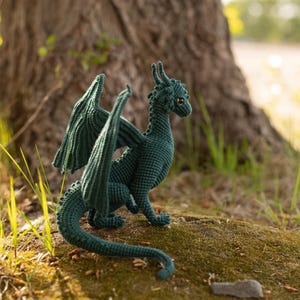 May include: A teal crocheted dragon figurine with outstretched wings, posed on a mossy surface. The handmade dragon has detailed scales, horns, and a long tail. The background features a tree trunk and natural light, highlighting the dragon's texture.