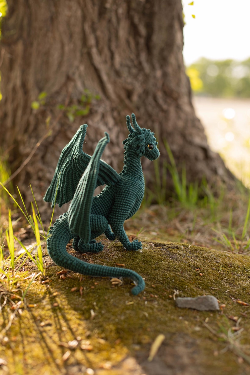 May include: A teal crocheted dragon figurine with outstretched wings, posed on a mossy surface. The handmade dragon has detailed scales, horns, and a long tail. The background features a tree trunk and natural light, highlighting the dragon's texture.