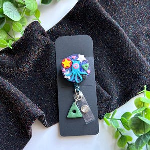 May include: A whimsical badge reel featuring a colorful space-themed design. The reel showcases a blue octopus, stars, and a planet, with a green triangle charm and a clear plastic clip. The background is a black card.