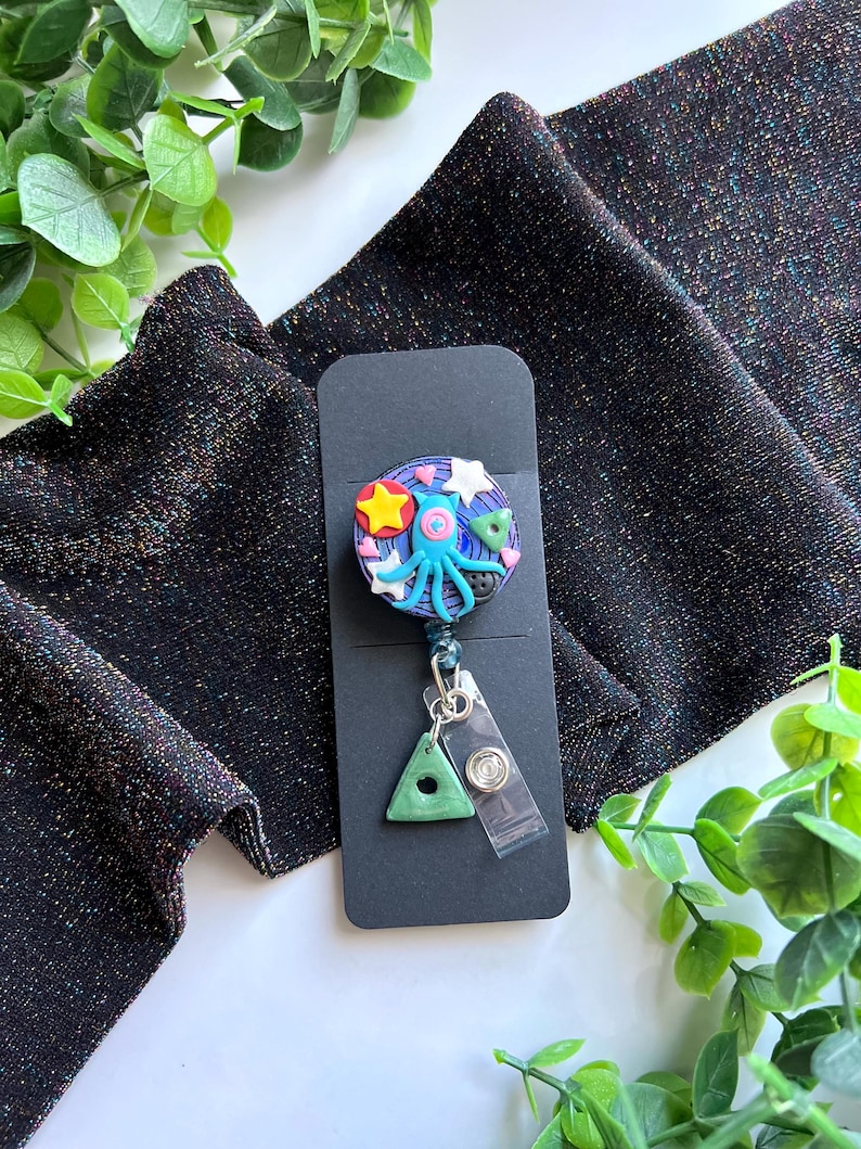 May include: A whimsical badge reel featuring a colorful space-themed design. The reel showcases a blue octopus, stars, and a planet, with a green triangle charm and a clear plastic clip. The background is a black card.