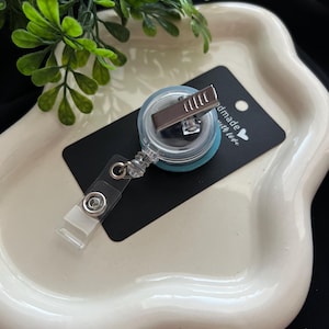 May include: A clear badge reel with a teal backing and a metal clip, resting on a black tag that reads "handmade with love." The reel is attached to a clear plastic strap. The items are on a white, wavy-edged tray.