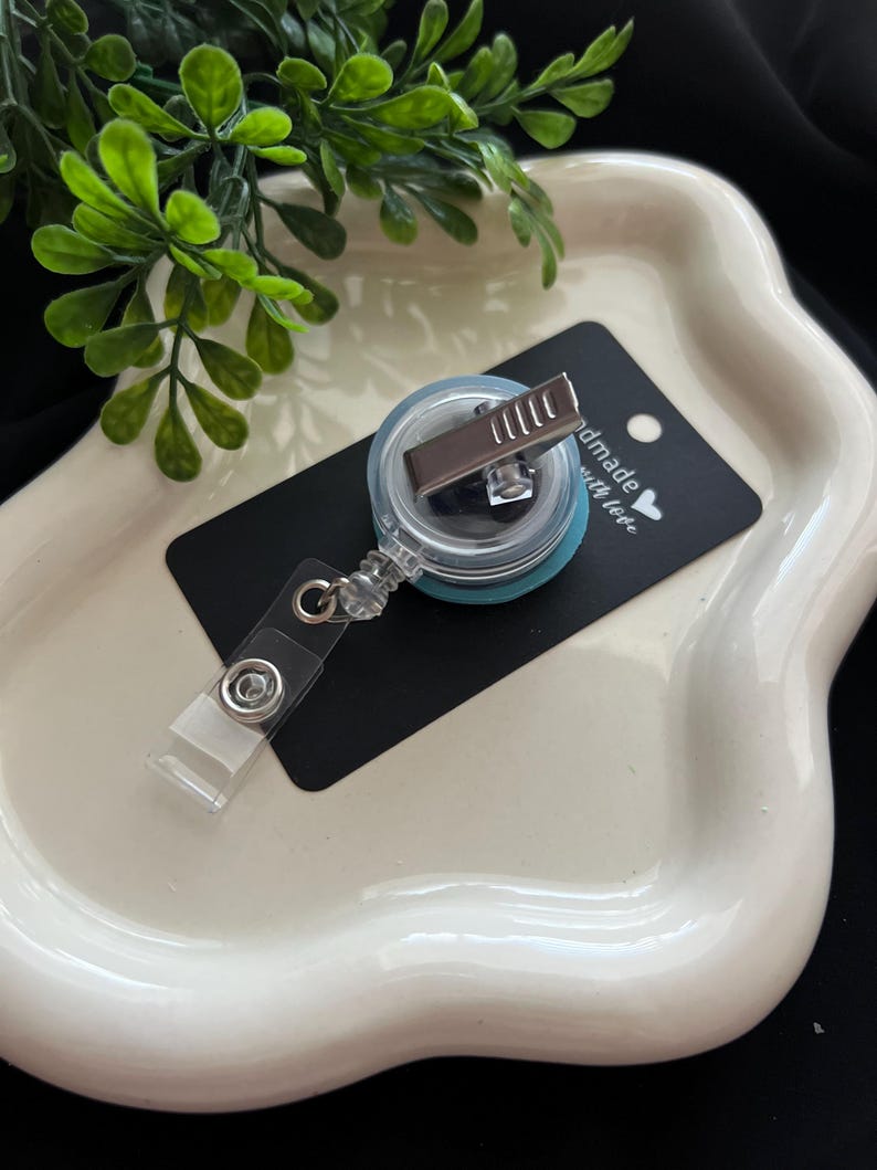 May include: A clear badge reel with a teal backing and a metal clip, resting on a black tag that reads "handmade with love." The reel is attached to a clear plastic strap. The items are on a white, wavy-edged tray.