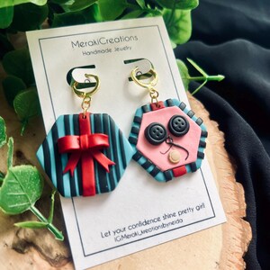 May include: A pair of handmade polymer clay earrings. One earring is a blue and black striped hexagon with a red bow. The other earring is a pink hexagon with black buttons and a white face.