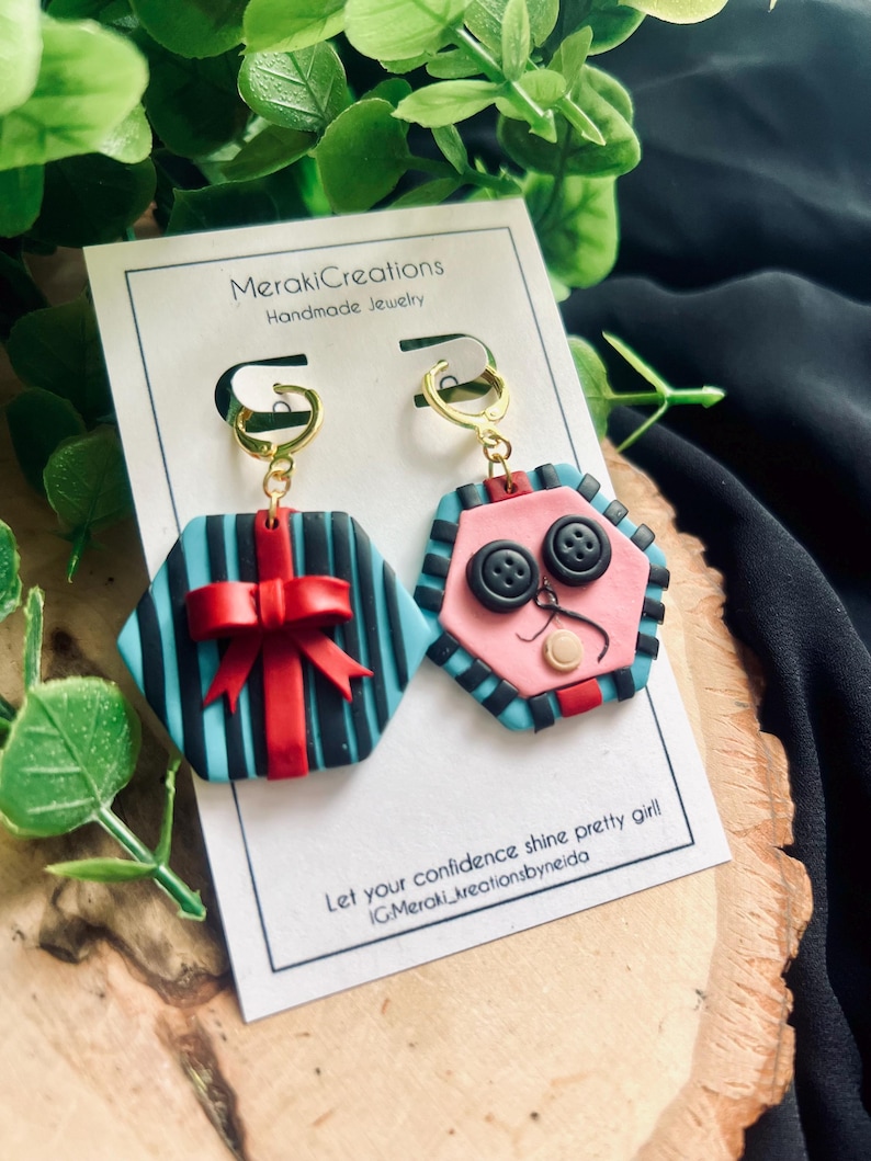 May include: A pair of handmade polymer clay earrings. One earring is a blue and black striped hexagon with a red bow. The other earring is a pink hexagon with black buttons and a white face.