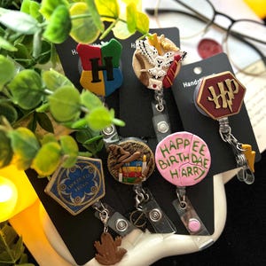 May include: A collection of themed retractable badge holders featuring various designs. The designs include a Hogwarts crest, an owl with a letter, and a pink badge with "Happee Birthdae Harry" text. Each badge holder has a clip.
