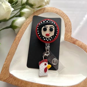 May include: A black and red badge reel featuring a cartoon face with black hair and red lips. The badge reel has a white and red chicken charm attached. The badge reel is on a black card.