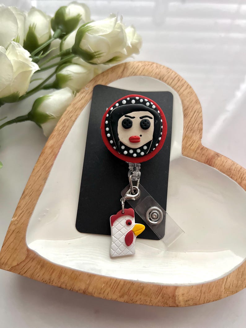 May include: A black and red badge reel featuring a cartoon face with black hair and red lips. The badge reel has a white and red chicken charm attached. The badge reel is on a black card.