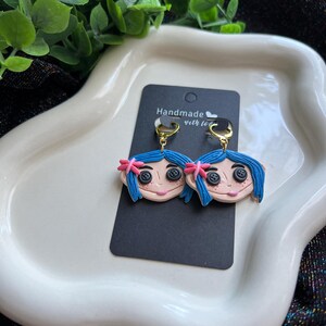 Pu&ograve; includere: Un paio di orecchini fatti a mano raffiguranti un personaggio dei cartoni animati con capelli blu, occhi a bottone e un fiocco rosa. Gli orecchini sono attaccati a ganci color oro e presentati su una carta nera con la scritta "Handmade with love".