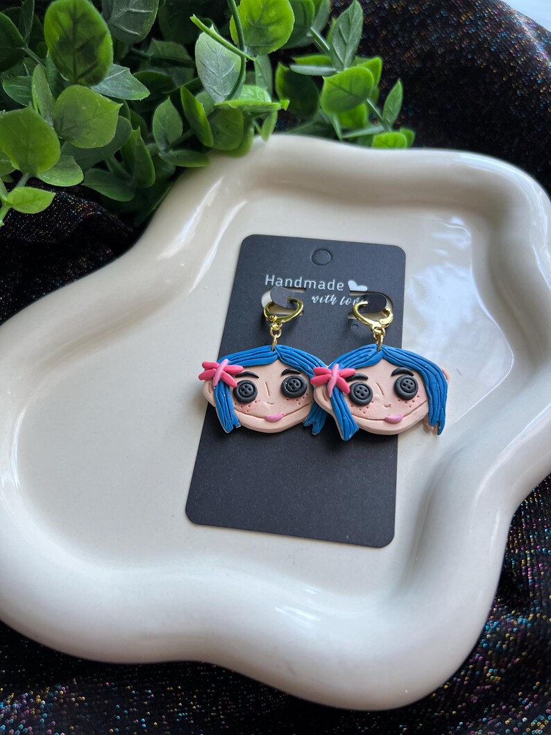 Pu&ograve; includere: Un paio di orecchini fatti a mano raffiguranti un personaggio dei cartoni animati con capelli blu, occhi a bottone e un fiocco rosa. Gli orecchini sono attaccati a ganci color oro e presentati su una carta nera con la scritta "Handmade with love".