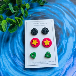 May include: A set of six handmade polymer clay earrings. The earrings are in the shape of a button, a flower, and a triangle. The button earrings are black with five holes, the flower earrings are red with a yellow center, and the triangle earrings are green with a hole in the center. The earrings are displayed on a white card with the text "MerakiCreations Handmade Jewelry" and "Let your confidence shine pretty girl IG:Meraki.creationsbyneida".