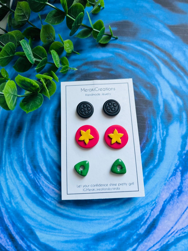 May include: A set of six handmade polymer clay earrings. The earrings are in the shape of a button, a flower, and a triangle. The button earrings are black with five holes, the flower earrings are red with a yellow center, and the triangle earrings are green with a hole in the center. The earrings are displayed on a white card with the text "MerakiCreations Handmade Jewelry" and "Let your confidence shine pretty girl IG:Meraki.creationsbyneida".