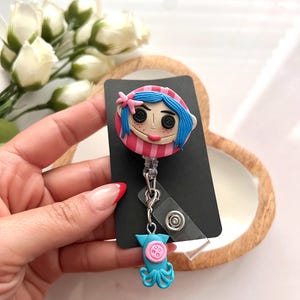 May include: A badge reel featuring a cartoon character with blue hair, button eyes, and a pink and red striped hat. The reel has a blue octopus charm with button details. The badge is attached to a black card.