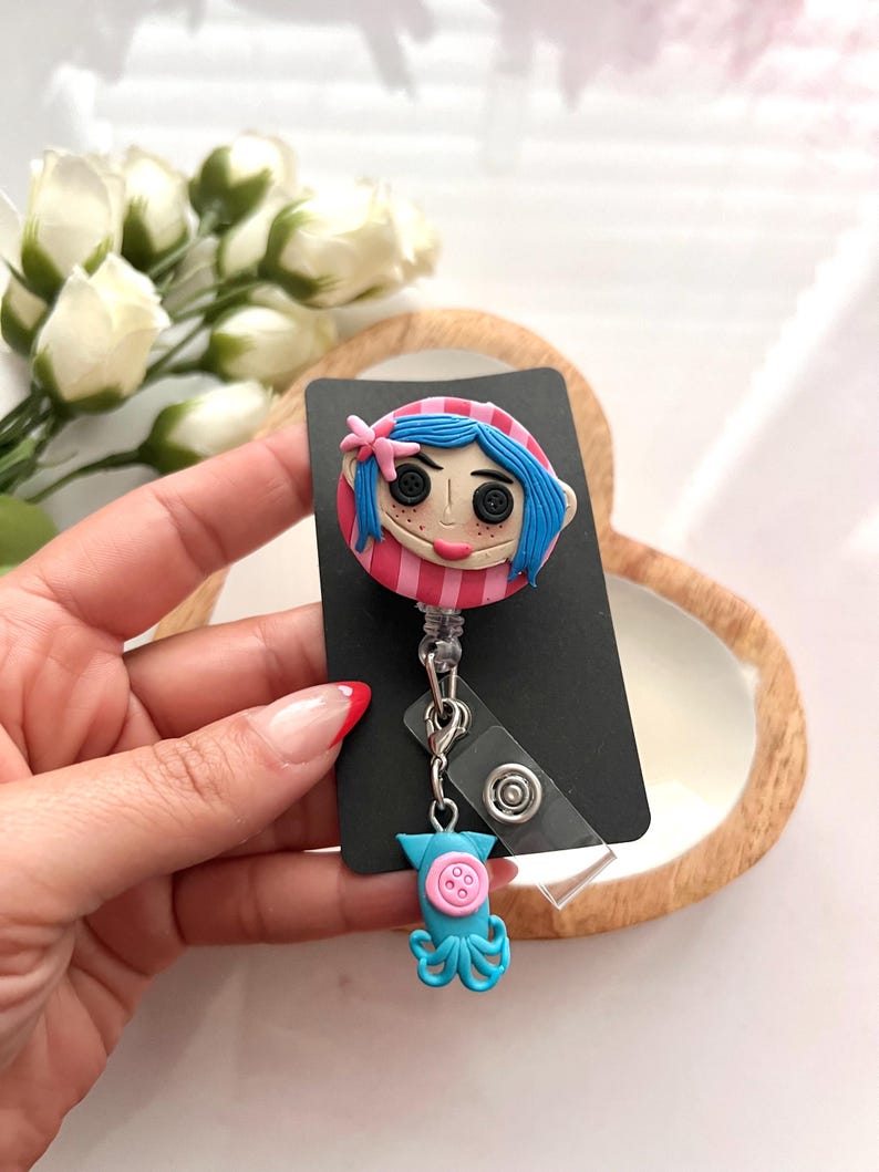 May include: A badge reel featuring a cartoon character with blue hair, button eyes, and a pink and red striped hat. The reel has a blue octopus charm with button details. The badge is attached to a black card.