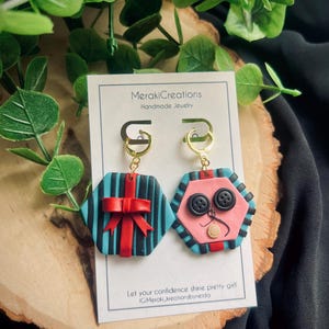 May include: A pair of handmade polymer clay earrings. One earring is a blue and black striped hexagon with a red bow. The other earring is a pink hexagon with black buttons and a white nose. The earrings are attached to gold hoops.
