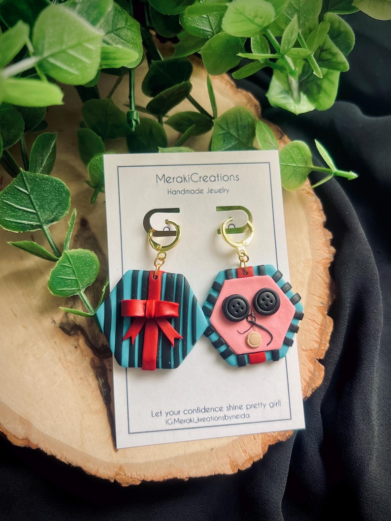 May include: A pair of handmade polymer clay earrings. One earring is a blue and black striped hexagon with a red bow. The other earring is a pink hexagon with black buttons and a white nose. The earrings are attached to gold hoops.