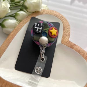 May include: A handmade badge reel with a black rectangular base. The badge reel features a colorful design with a gear shift, a yellow star, and a white sphere. The reel has a clear plastic strap and a metal clip.