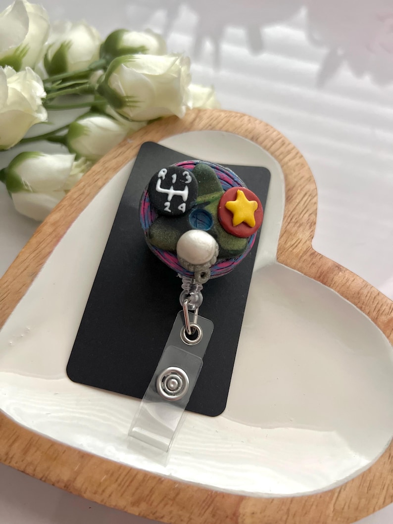 May include: A handmade badge reel with a black rectangular base. The badge reel features a colorful design with a gear shift, a yellow star, and a white sphere. The reel has a clear plastic strap and a metal clip.