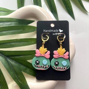 May include: A pair of green and pink Stitch earrings with gold hooks. The earrings are handmade with love and feature a pink bow and a blue button.