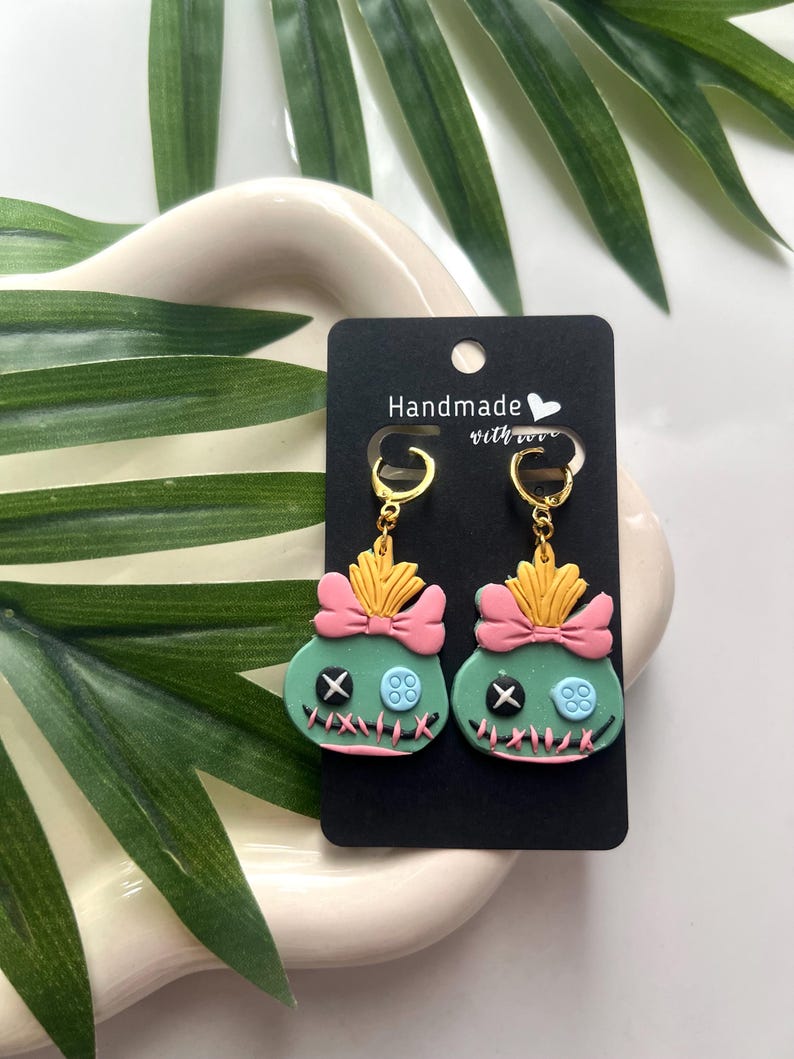 May include: A pair of green and pink Stitch earrings with gold hooks. The earrings are handmade with love and feature a pink bow and a blue button.