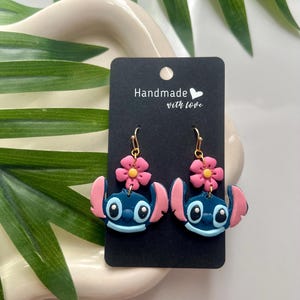 May include: A pair of blue Stitch character earrings with pink flower accents. The earrings are handmade with love and are displayed on a black card with the text "Handmade with love".
