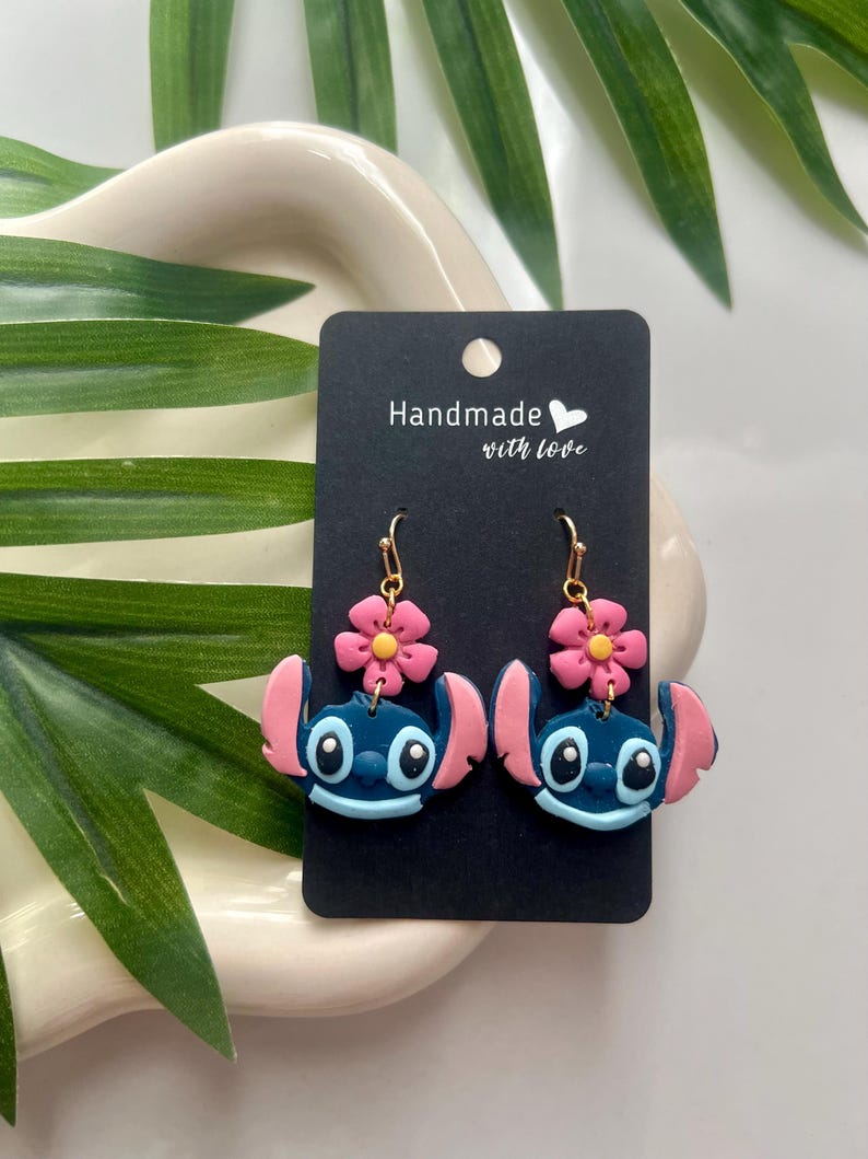 May include: A pair of blue Stitch character earrings with pink flower accents. The earrings are handmade with love and are displayed on a black card with the text "Handmade with love".