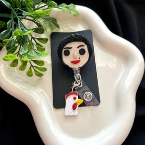 May include: A black and white badge reel featuring a cartoon character with black hair, red lips, and button eyes. A small chicken charm hangs below. The reel is on a black card, set against a white, wavy dish with green foliage.