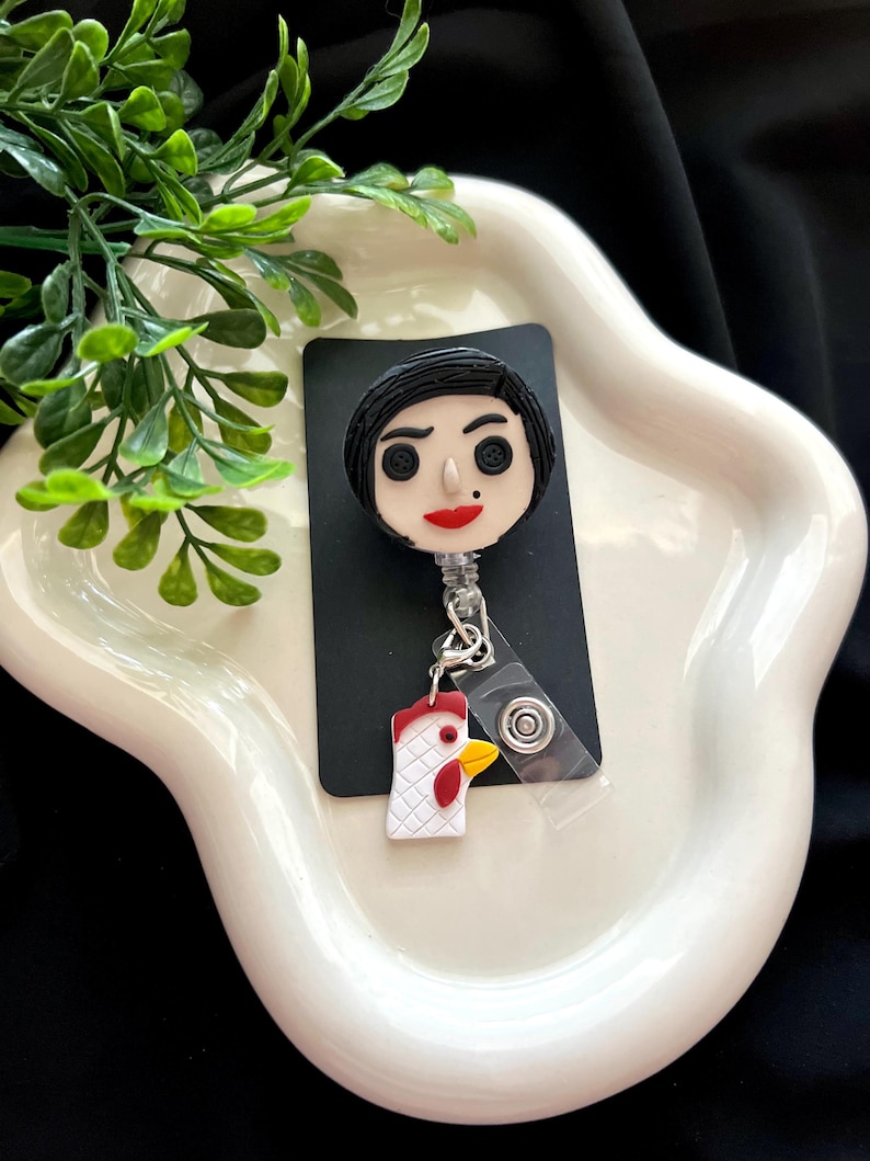 May include: A black and white badge reel featuring a cartoon character with black hair, red lips, and button eyes. A small chicken charm hangs below. The reel is on a black card, set against a white, wavy dish with green foliage.