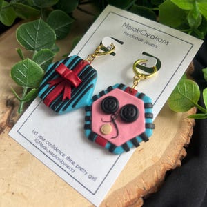 May include: A pair of handmade polymer clay earrings. One earring is shaped like a gift box with a red bow and blue and black stripes. The other earring is shaped like a face with pink skin, black buttons for eyes, and a black and blue striped border.