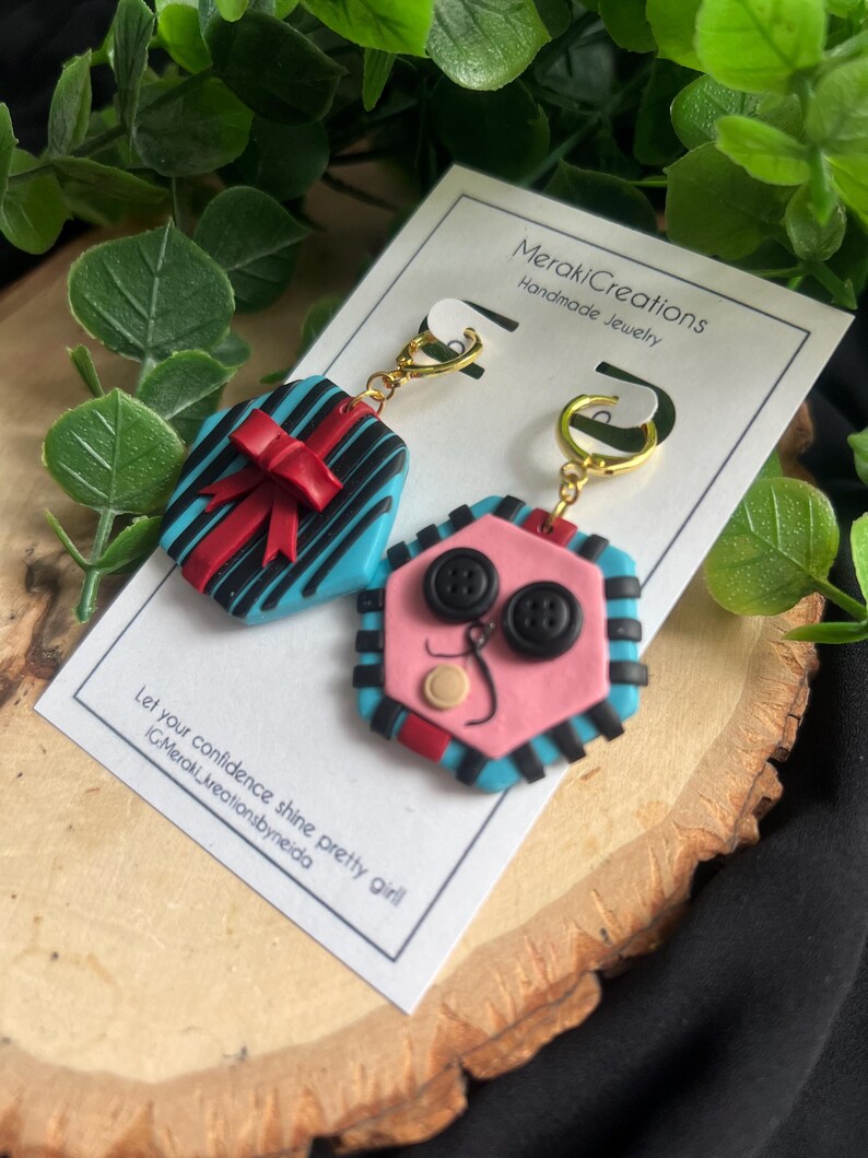 May include: A pair of handmade polymer clay earrings. One earring is shaped like a gift box with a red bow and blue and black stripes. The other earring is shaped like a face with pink skin, black buttons for eyes, and a black and blue striped border.