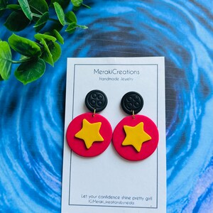 May include: A pair of handmade polymer clay earrings featuring a red circle with a yellow star and a black button charm. The earrings are displayed on a white card with the text "Meraki Creations Handmade Jewelry" and "Let your confidence shine pretty girl IG:Meraki_kreationsbyneida".