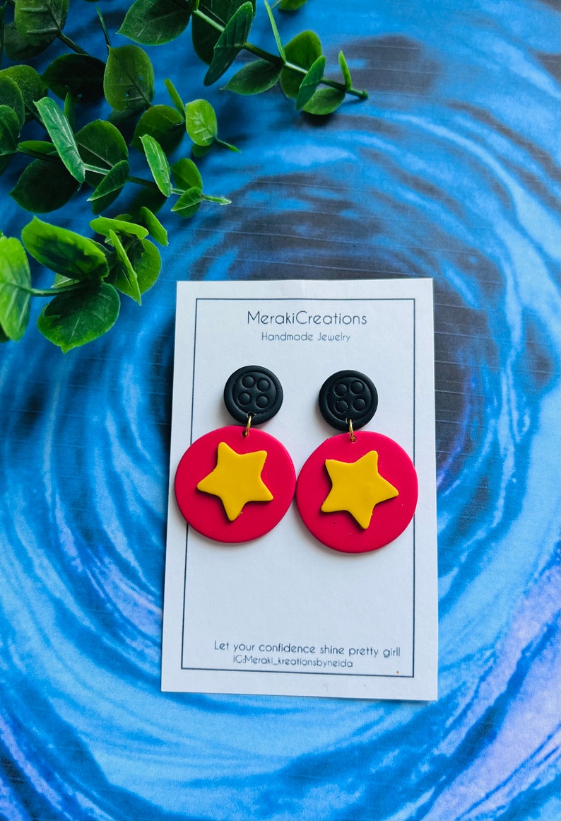 May include: A pair of handmade polymer clay earrings featuring a red circle with a yellow star and a black button charm. The earrings are displayed on a white card with the text "Meraki Creations Handmade Jewelry" and "Let your confidence shine pretty girl IG:Meraki_kreationsbyneida".