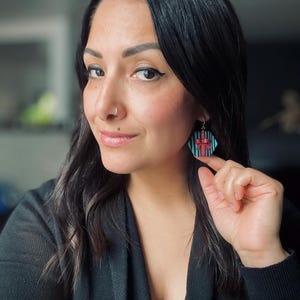 May include: A woman wearing a black sweater with a pair of blue and red geometric earrings. The earrings are shaped like a gift box with a red bow.