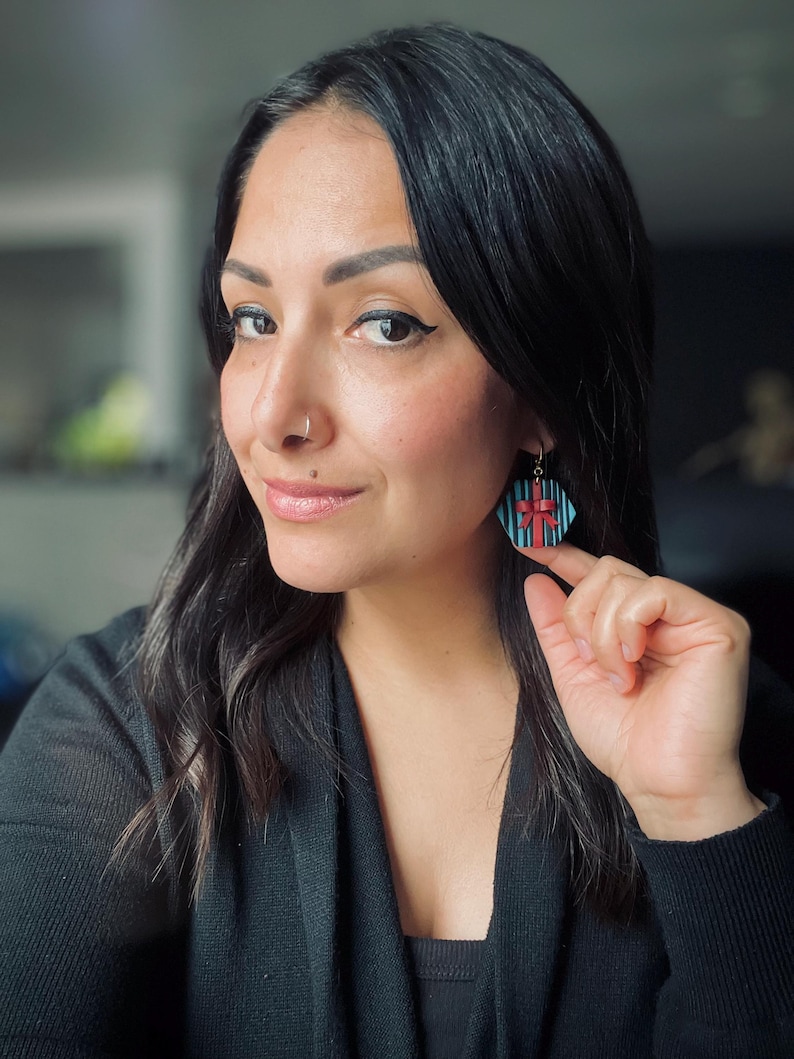 May include: A woman wearing a black sweater with a pair of blue and red geometric earrings. The earrings are shaped like a gift box with a red bow.