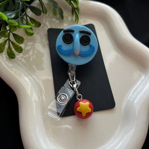 May include: A light blue badge reel featuring a cartoon face with button eyes and a blue mustache. A red ball with a yellow star charm hangs from the reel. The reel is attached to a clear plastic clip.