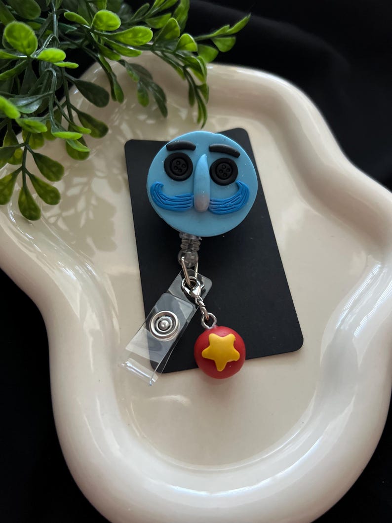 May include: A light blue badge reel featuring a cartoon face with button eyes and a blue mustache. A red ball with a yellow star charm hangs from the reel. The reel is attached to a clear plastic clip.