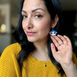 Pu&ograve; includere: Orecchini con personaggio dei cartoni animati blu e bianchi con ganci color oro. Gli orecchini presentano un personaggio con i capelli blu, un fiocco rosa e un viso sorridente. Gli orecchini pendono dall'orecchio.