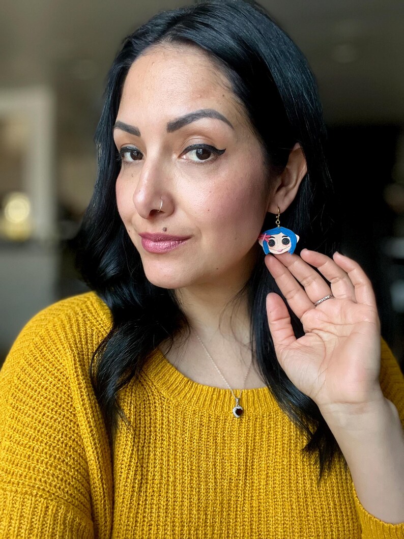 Pu&ograve; includere: Orecchini con personaggio dei cartoni animati blu e bianchi con ganci color oro. Gli orecchini presentano un personaggio con i capelli blu, un fiocco rosa e un viso sorridente. Gli orecchini pendono dall'orecchio.