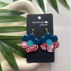 May include: A pair of teal blue and red floral earrings with white leaf designs. The earrings are handmade with love.
