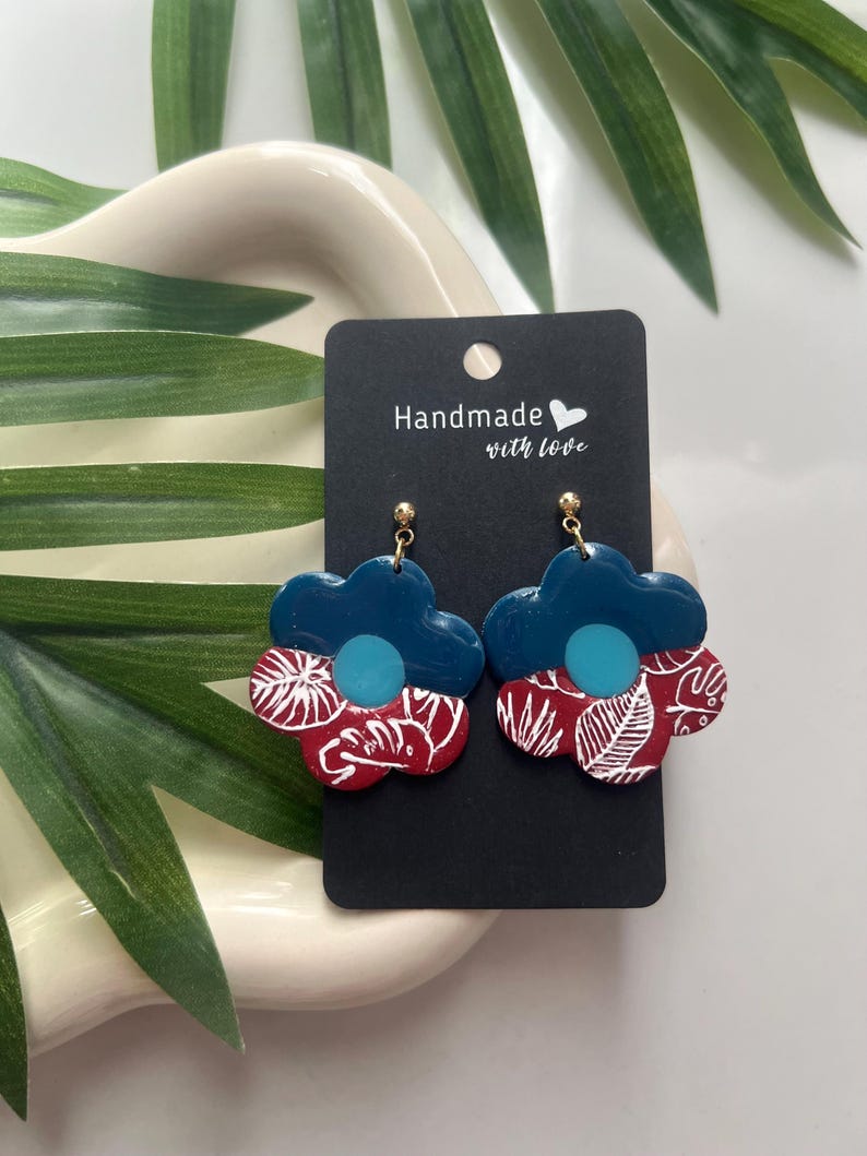 May include: A pair of teal blue and red floral earrings with white leaf designs. The earrings are handmade with love.