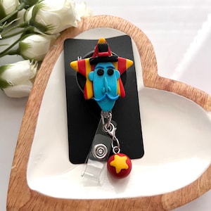May include: A whimsical badge reel featuring a blue-faced character with a mustache, a star-shaped head, and a red and yellow hat. A red ball with a yellow star charm hangs below. The reel is attached to a black card.