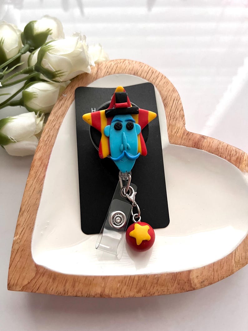 May include: A whimsical badge reel featuring a blue-faced character with a mustache, a star-shaped head, and a red and yellow hat. A red ball with a yellow star charm hangs below. The reel is attached to a black card.
